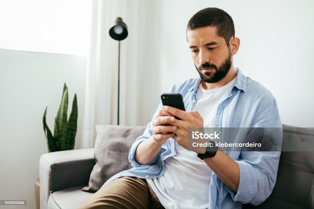Young brazilian man sitting on sofa using smartphone Men Stock Photo