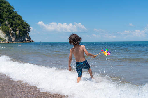 Boy playing in the waves crashing on the shoreline. He have a colorful weather vane toy in his hand. Shot with a full frame camera on a sunny summer day.