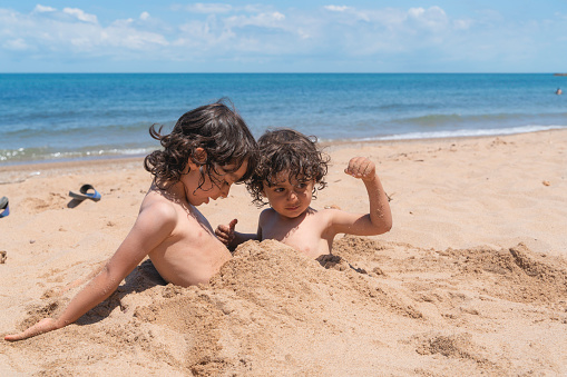 The brothers are sunbathing on the beach, buried in the sand. Having fun on the beach on a sunny and hot summer day. \nShot with a full frame camera.