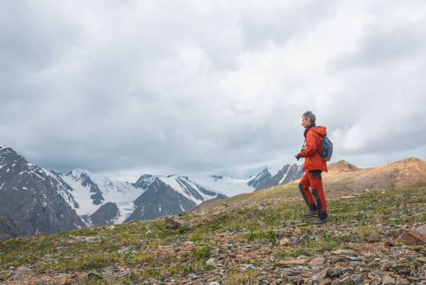 istock Man in red with camera walks with view to large snowy mountain range with sharp tops under gray cloudy sky. Dramatic landscape with tourist in high mountains. Snow pointy peaks at changeable weather. 1410535510