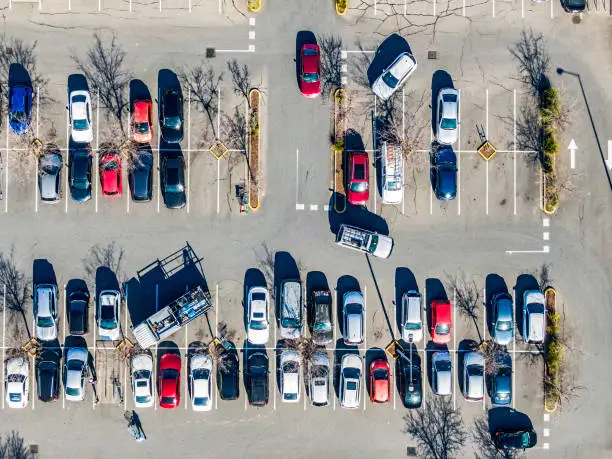 Aerial view directly above suburban shopping centre carpark; trade truck parked diagonally across 3 spaces; cars arriving and exiting parking spaces, woman returning shopping trolley, mostly red, blue, white and grey cars, direction arrow, bare trees, shadows.