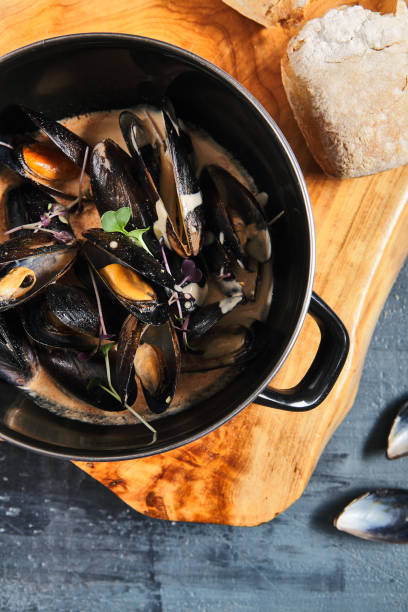 Mussel stew pot with creamy sauce, baguette on stone table. Mussels soup pot contemporary style. Aesthetic composition with seafood. Gastronomy mussel pot dinner concept. Stone table and wooden tray olla de guiso de mejillones con salsa cremosa, baguette sobre mesa de piedra. olla de sopa de mejillones al estilo contemporáneo. composición estética con mariscos. concepto de cena de mejillón de gastronomía. mesa de piedra y bandeja de madera.