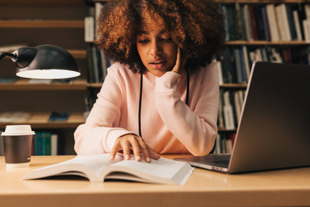 African American girl reading from book in library while preparing exams at evening. Student sitting at desk. stock photo