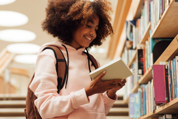 Smiling girl holding a book in library while standing at bookshelf stock photo