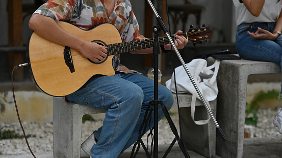 Asian male artist dressed like a hipster playing the guitar next to a microphone standing in the park. Music and abstract concepts.