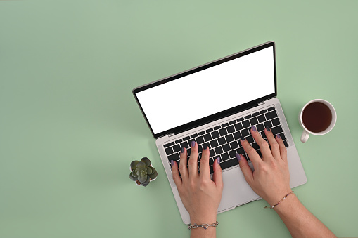 Male hands with colored nails and bracelets using a laptop computer with an empty white screen between a cactus and a cup of coffee on the light green background. Flat lay composition.