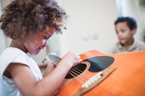 African American schoolgirl with acoustic guitar looking at paper with musical notes while playing instrument against classmates