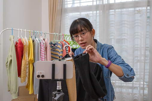 Young Asian woman selling online clothing using smart phone via social media at home, Internet or wireless technology, E-commerce.