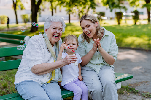 A happy smiling woman with senior grandmother and little daughter blowing soap bubbles at park, family, generation and people concept.