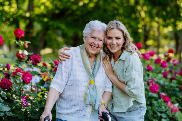 retrato de nieta adulta con abuela mayor a pie en el parque, con rosas al fondo - granddaughter fotografías e imágenes de stock