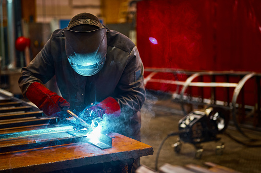 Skilled worker in uniform and protective mask welds manually metal part in shielding gases at production plant close side view