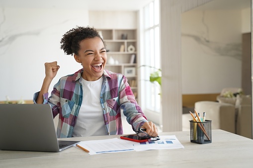 Exam is passed successfully. Happy excellent african american girl is winner. Teenage girl is sitting in front of computer and celebrating her victory. Remote study, distance learning online at home.