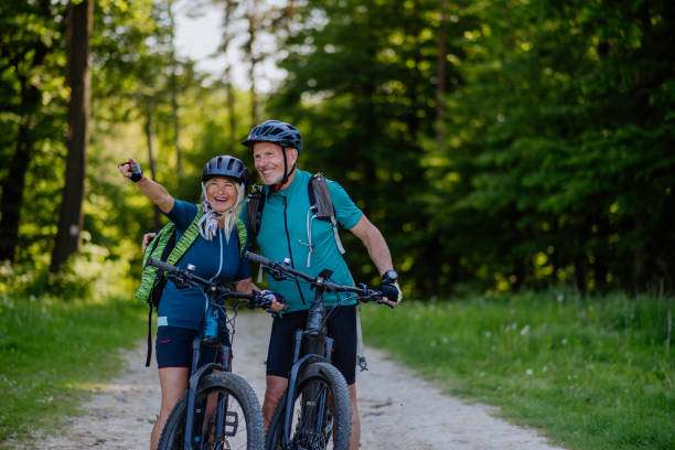 portret aktywnej pary seniorów jeżdżącej na rowerach w parku letnim, stojącej na ścieżce i patrzącej w kamerę - cycling senior adult sports helmet men zdjęcia i obrazy z banku zdjęć