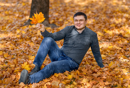 portrait of a man, he is sitting in a glade with yellow maple leaves, a bright sunny day in an autumn park
