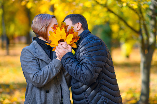 portrait of a romantic couple in an autumn city park, a man and a woman posing, they cover their faces with yellow leaves and kiss, a bright sunny day