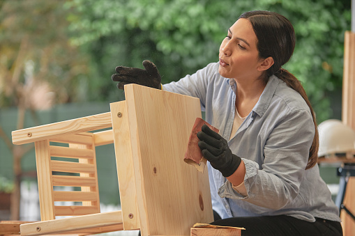 Diligent female carpenter preparing a table to be painted or stained. Young woodworker or craftswoman making furniture as hobby or to sell. Serious woman sanding restored wood with sandpaper outside.