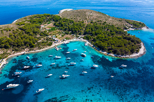Beautiful Marinkovac island, part of Pakleni islands, with its amazing beach and incredible shades of blue and emerald colors with many anchored boats seen from a drone perspective during summer season in Croatia.