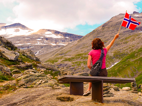Tourist woman holds norwegian flag, enjoying nature landscape from Trollstigen viewpoint area. Trolls Path mountain road in Norway. Touristic attraction.