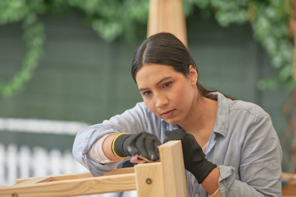istock Female carpenter preparing a table to be painted or stained for DIY project. Young woodworker or craftswoman making furniture as a hobby. Serious woman sanding restored wood with sandpaper outside. 1410619744