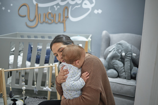 Mother hugging her baby inside a nursery room with a traditional name mural painted on the wall. Single muslim female holding her baby in a modern grey child bed room with a crib and toys.