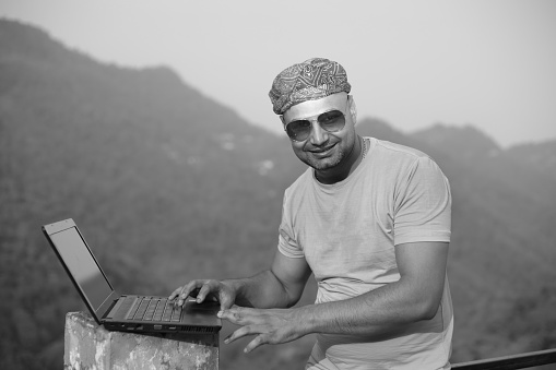 Outdoor image of young Indian  man working on laptop in outdoor with the railing by the side of road in hilly area. He is in casual dress. One person, Waist up and selective focus. Shoot location Naina Devi, ( in mountain) Himachal Pradesh, India.