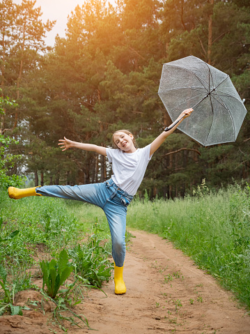 Teenager girl with transparent umbrella poses raising leg against green rainy forest. Smiling young lady dressed in white T-shirt jeans and yellow rubber boots