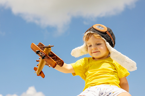 Happy child boy playing with wooden toy airplane on blue sky background with clouds.