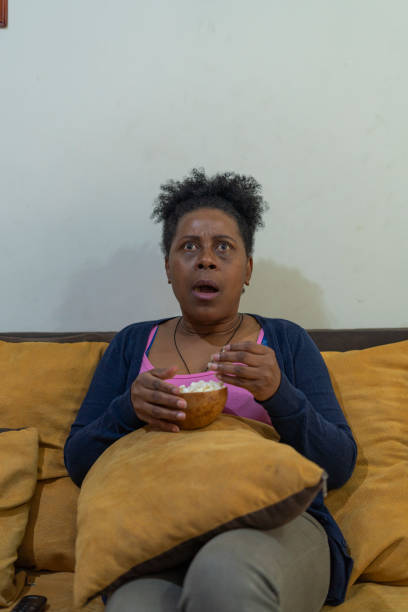 mujer negra viendo el capítulo de su serie favorita en su televisión sentada en los muebles de su casa comiendo palomitas de maíz - chapter one fotografías e imágenes de stock