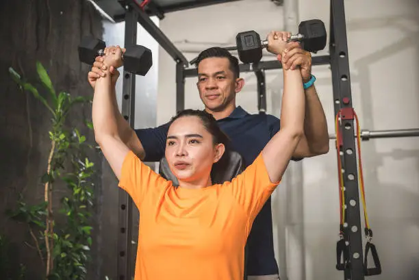 An instructor spots his female client during a set of dumbbell shoulder presses. Assisting a lift for proper form and safety. At a home gym.