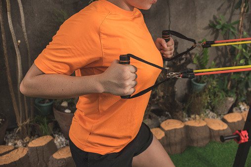 An anonymous woman of intermediate fitness level does standing back rows with a resistance band at a home gym.