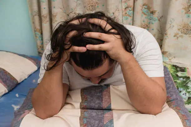 Photo of A depressed man in his 30s sitting on the bed and grasping his hair. Bad news sinking in and contemplating.