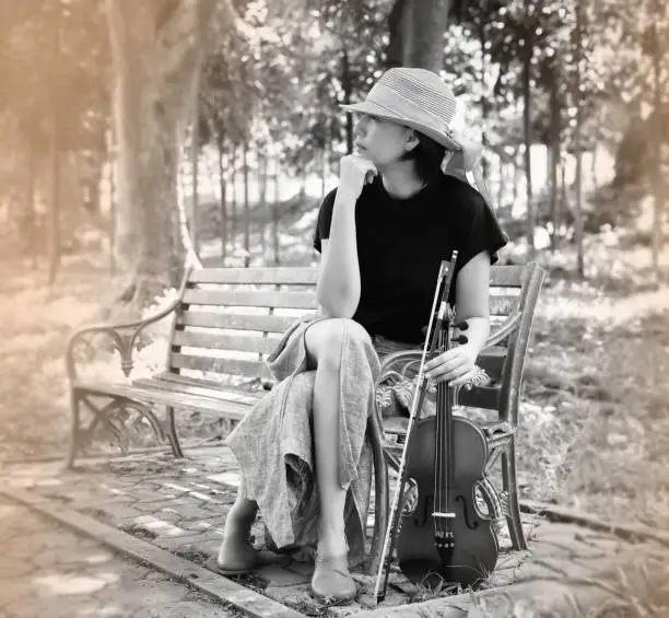 Photo of Woman wearing woven hat sitting on old and rust bench,holding violin and bow in hand,in a park