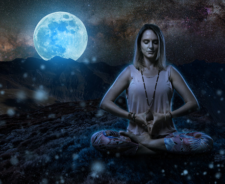 Woman yoga practitioner meditating during the moon ritual in the mountains, blue toned at midnight