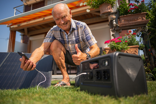 Smiling senior man showing thumb up, very happy to charge his mobile phone from portable solar panels and generator, set up outdoors in his frontyard garden on sunny summer day