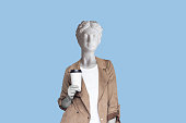 Young successful business woman headed by antique statue holds a white paper cup pf tea or coffee