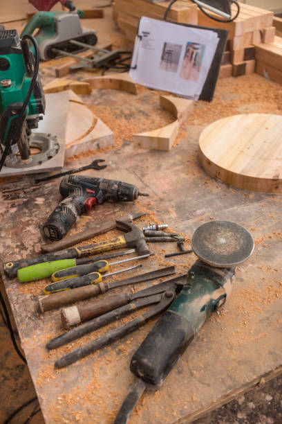 istock Woodworking tools on a table filled with sawdust. Drills, a hammer, screwdrivers, chisels and an angle grinder. A finished circle slab with reference drawing for a stool design at the back. 1410657915