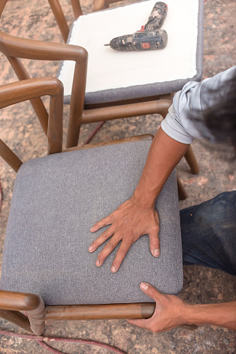 An upholsterer fits a cushioned pad into a wooden chair. At an upholstery, furniture, or woodworking shop.