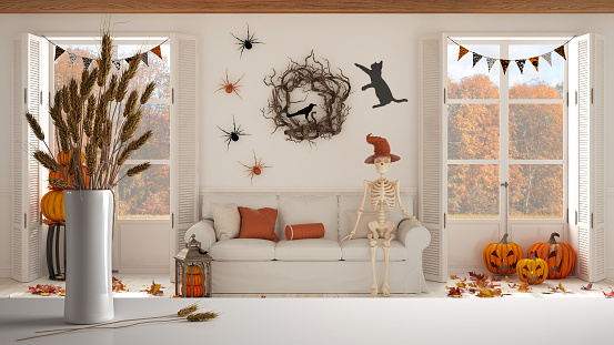 White table top or shelf with straws, dry plants, ornament, ears, sheaf, branch in vase, over Halloween living room with fireplace and autumnal landscape, classic interior design