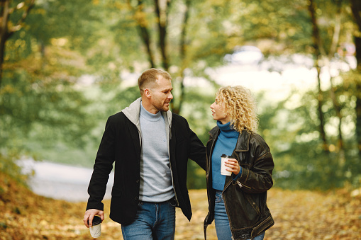 Blonde curly woman and man holding hands in autumn forest and hugging. Couple wearing leather jackets and blue sweaters. Photo of romantic couple in a forest.