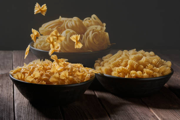 istock Three deep bowls with uncooked pasta on a wooden table. Assortment of raw macaroni. Falling paste, levitation effect 1410667940
