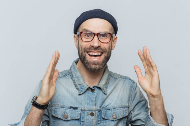 istock Handsome happy man raises hands with excitment, has overjoyed expression, looks with delighted face, has thick beard and mustache. Glad man in eyewear poses against white background, has fun 1410673414
