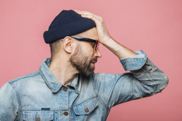 istock Sideways portrait of bearded male has regretful expression, keeps hand on forehead, looks desperately, isolated over pink background. Stressful unshaven man in glasses has terrible headache. 1410673452