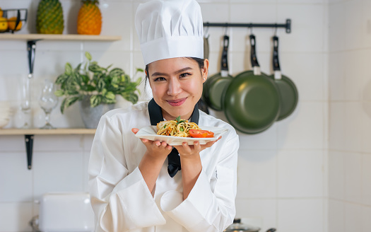 Portrait beautiful Asian professional female chef wearing white uniform, hat, showing plate of spaghetti, cooking in kitchen, making surprising face with happiness. Restaurant, Food Concept