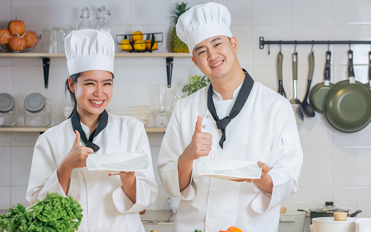 Two Asian professional couple chef wearing white uniform and hat, showing blank space of plates, thumn up, cooking in kitchen together, smiling with happiness, confidence. Food Concept