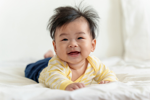 Portrait of Asian cute little baby boy relaxing in bed, smiling and looking at camera. Happy Adorable newborn baby having tummy time on bed in white sunny bedroom.