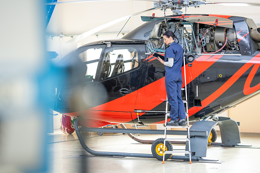 Female aircraft mechanic in uniform standing on ladder near jet, back view. Engineer working in air helicopter hangar and looking in tablet, wide shot