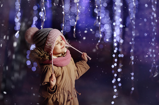 A child girl in a winter jacket and a hat stands on the street among beautiful New Year's garlands. Enjoying the New Year mood.