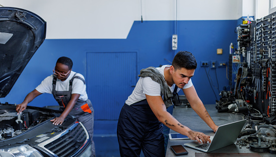 A young Caucasian male mechanic is typing on his laptop, with his young African American colleague fixing a car behind him.