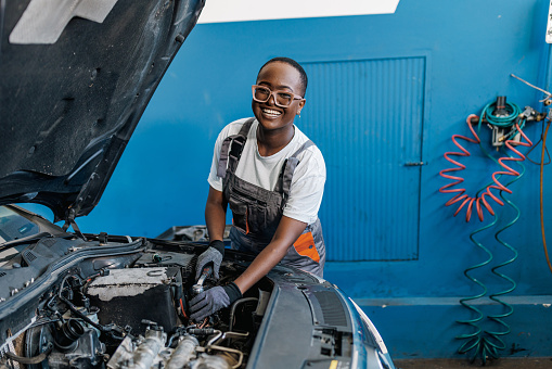 A young African American female car mechanic is repairing a car and looking at the camera with a big smile on her face.