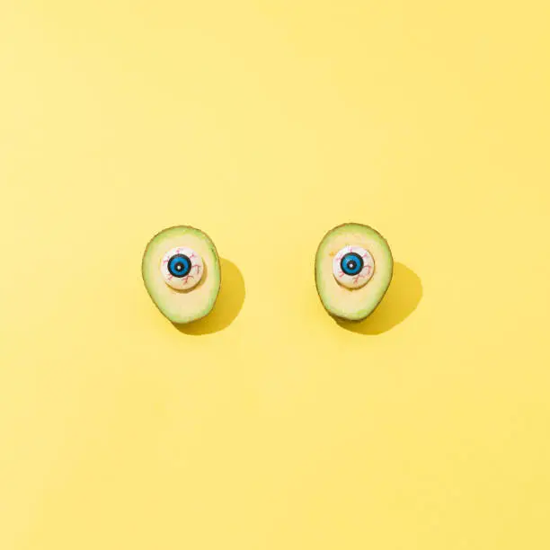 Sliced avocado with plastic eyes instead of stone on yellow background. Happy halloween concept with copy space.  Square ratio.